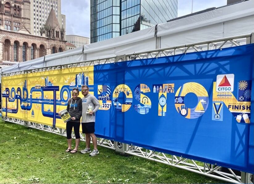 Showing the CITGO Road to Boston graphic MDR created for the Boston Marathon hanging on a fence with two people posing in front of it.